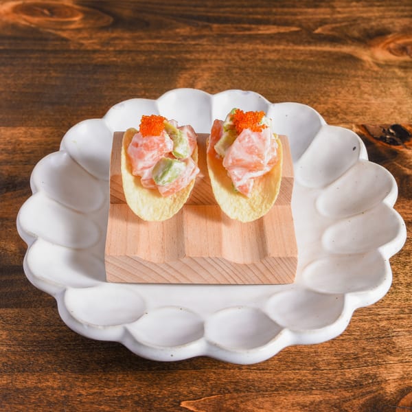 Two shrimp and avocado appetizers topped with orange roe, served on a wooden board atop a scalloped white plate.