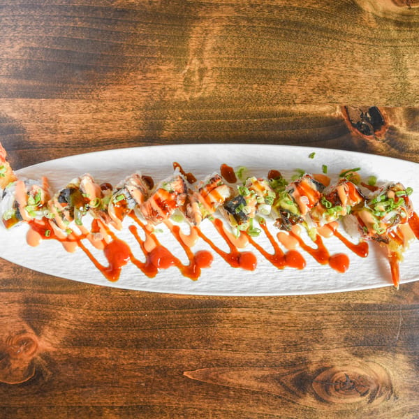 Shrimp sushi roll topped with spicy mayo, eel sauce, and scallions on a white platter over a wooden table.