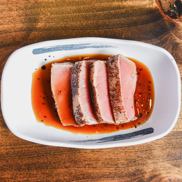 Seared tuna slices with peppered edges served in a glossy reddish-brown sauce on a white plate.