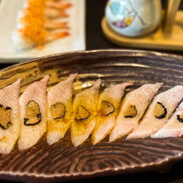 Thinly sliced fish sashimi with truffle shavings and sauce on a decorative brown plate.