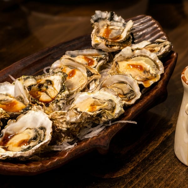 Fresh oysters on a brown platter with sauce, next to a ceramic fish-decorated soy sauce dispenser.