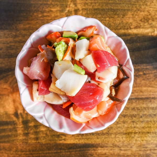 Colorful seafood salad with raw fish, shellfish, avocado, and vegetables in a scalloped bowl on a wooden table.