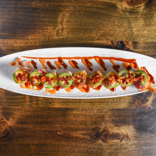 Sushi roll topped with jalapeño slices, spicy sauce, and shrimp, served on an elongated white plate over a wooden table.